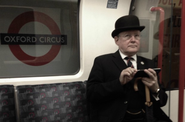 A man in bowler hat and 3-piece suit on the Tube using a smart phone.